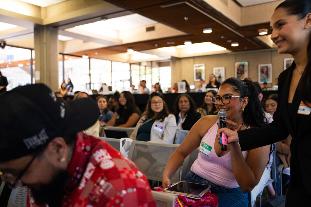A young Latina woman smiles while speaking into a microphone. The microphone is held by another young Latina woman. Other attendees are seen seated in the background.